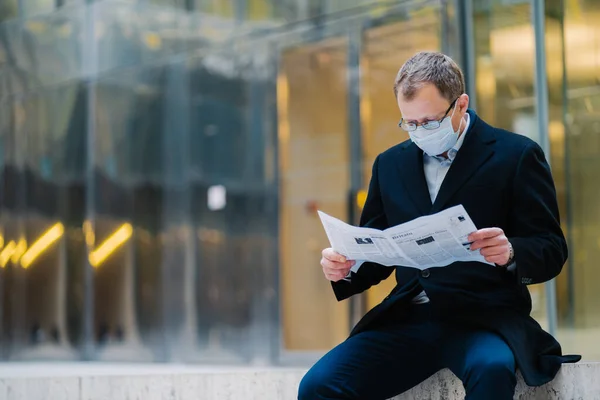 Outdoor shot of serious man boss takes break after walking, reads newspaper, wears spectacles for good vision, medical mask to protect himself from viruses, finds out news about spreading coronavirus