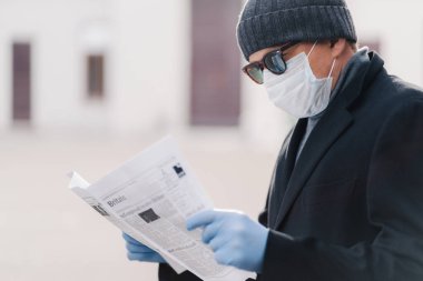 Koronavirüs için karantina. Yetişkin adamın koruyucu maske taktığı ve tek kullanımlık eldiven taktığı, yeni gazete okuduğu ve salgın hastalıkla ilgili haberleri öğrendiği yatay bir görüntü. Enfeksiyon önleme kavramı