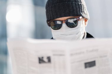Yetişkin bir adamın güneş gözlüğü, şapka ve steril sağlık maskesi takıp, gazete okurken, dünyadaki durumu öğrenirken, koronavirüs yayılıp, bulanık arka planda poz verirken yakın plan çekimleri.