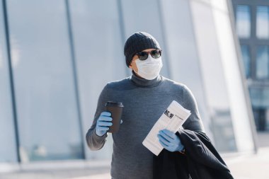 Genç bir adamın steril maske ve lastik tıbbi eldiven giydiği, bulaşıcı hastalıklar sırasında kenarda yürüdüğü, kahve içtiği, gazete katladığı görüntüsü. Coronavirus önleme kavramı