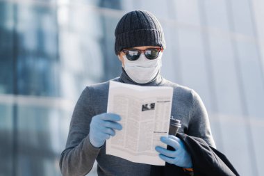 Genç adam Coronavirus belirtileri hakkında makale okuyor, koruyucu maske ve lastik eldiven takıyor, sokakta kahve ile poz veriyor, kurallara uyuyor veya karantinaya alınıyor. Covid-19 koruma kavramı