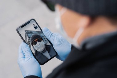 Tanınmayan bir adam dışarıda görüntülü konuşma yapıyor, cep telefonunu koruyucu lastik eldivenlerle tutuyor, koronavirüs salgını yayıldığı için koruyucu maske takıyor, arkadaşlarıyla konuşuyor, haberleri tartışıyor.
