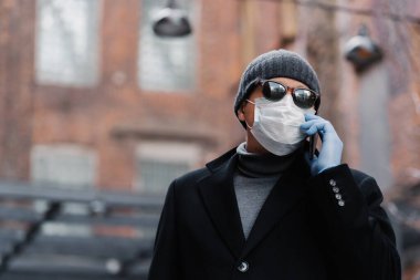 Hasta adam virüs salgını sırasında koruyucu maske takıyor, arkadaşıyla telefonda konuşuyor, siyah ceket ve güneş gözlüğü takıyor, dışarıda poz veriyor. Sağlığınızı koruyun. Coronavirus, panik konsepti