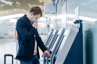Shot of man passenger poses near check in desk in airport, has self service, wears medical mask to avoid spreading respiratory disease during coronavirus outbreak, travels and going to fly abroad clipart