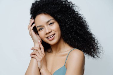 Güzellik, cilt bakımı konsepti. Afro saçlı güzel genç bir kadın sağlıklı cilde sahip, saçlara dokunuyor, kameraya olumlu bakıyor, çıplak omuzlar gösteriyor, beyaz arka planda izole edilmiş..