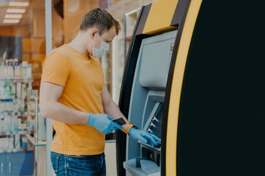 Man wears protective mask and medical gloves, uses ATM machine, prevents from virus pandemic, introduces pin code. Safe money withdrawal during coronavirus outbreak. Payment, influenza concept clipart