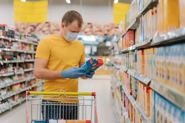 Young man stands in big supermarket near cart, reads label of product, wears yellow t shirt, medical mask and rubber gloves, prevents from coronavirus. Purchase of products during quarantine