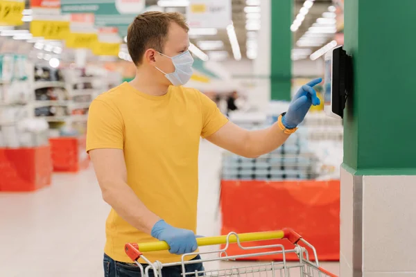 Photo of male consumer uses touchscreen in shop, checks price, poses with shopping cart, wears disposable mask and gloves, pays with self checkout. Coronavirus pandemic, safety, protective measures