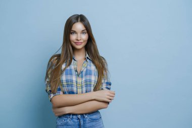 Uzun düz saçlı, kareli gömlekli, kot pantolonlu, ellerini çapraz tutan, temiz ciltli, minimum makyajlı, pozitif duygularını ifade eden, fotoğraf çekiminden hoşlanan çekici genç bir kadının yatay görüntüsü.