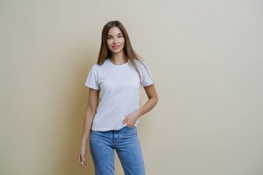 Stüdyo çekimlerinde oldukça zayıf bir kadın elini cebine atıyor, rahat bir tişört ve kot giyiyor, rahat bir poz veriyor, kendine güvenen kameraya bakıyor, bej arka planda müşteriyle gelişigüzel konuşuyor.