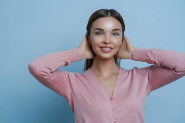 Mükemmel, temiz tenli, dişlek, nazik gülüşlü, ellerini saçından ayırmayan, pembe kazak giyen, stüdyoda fotoğraf çeken, mavi arka planda poz veren güzel bir kadının portresi. Kozmetik ve güzellik kavramı