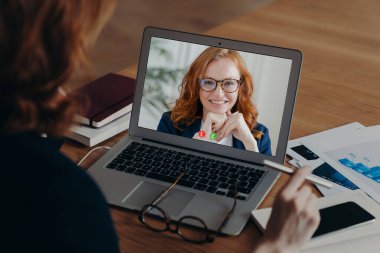 Kadın serbest çalışanın çalışanıyla video görüşmeleri, iş stratejisi hakkında görüşmeler. Gözlüklü, gülümseyen kızıl kadının dizüstü bilgisayar görüntüsü. Yeni teknoloji kullanımı. Mesafe çalışıyor