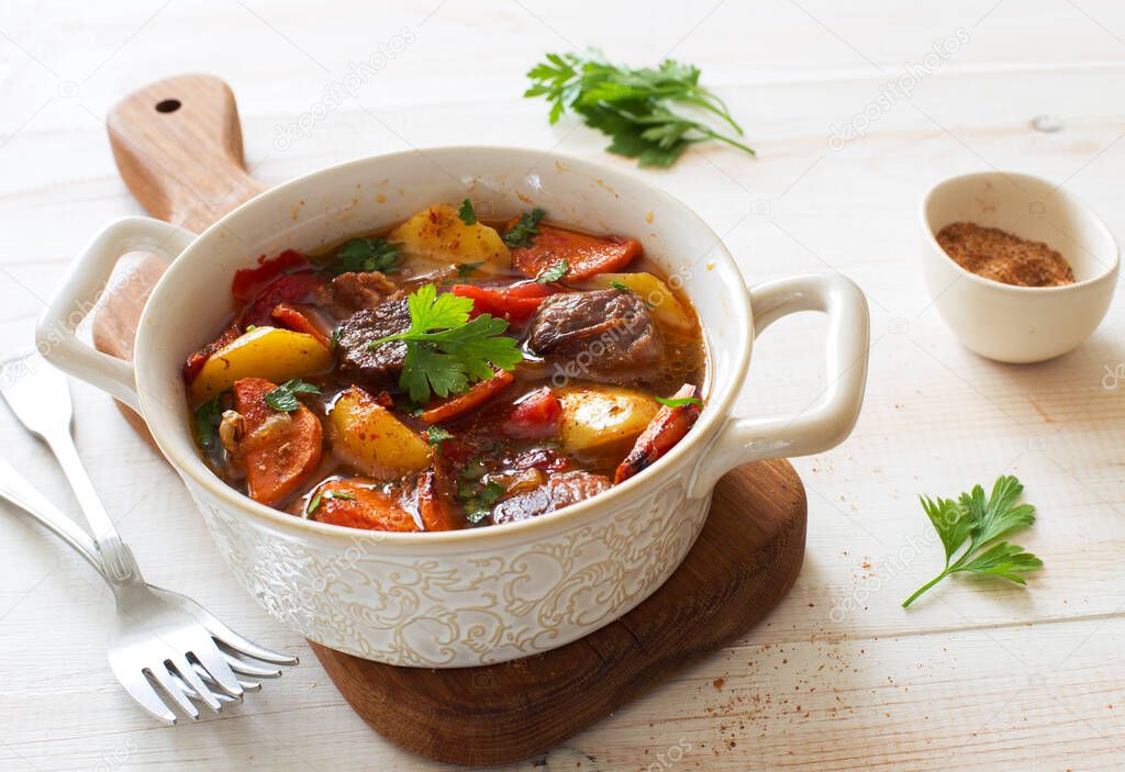 Beef stewed with vegetables( potatoes, carrots, tomatoes, onions) in a clay pot on a white wooden background close up.