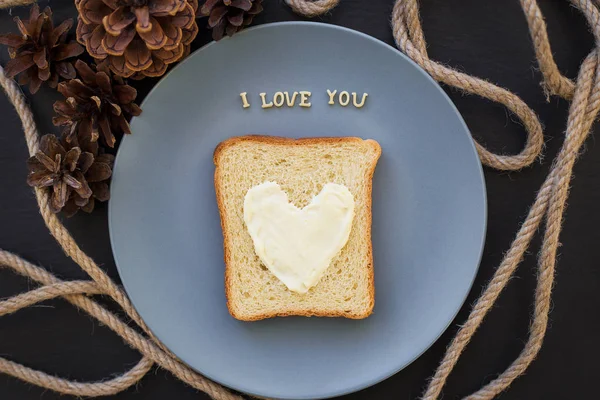 Panino per la prima colazione a forma di cuore con formaggio su un piatto blu e sfondo nero con coni iscrizione ti amo — Foto Stock