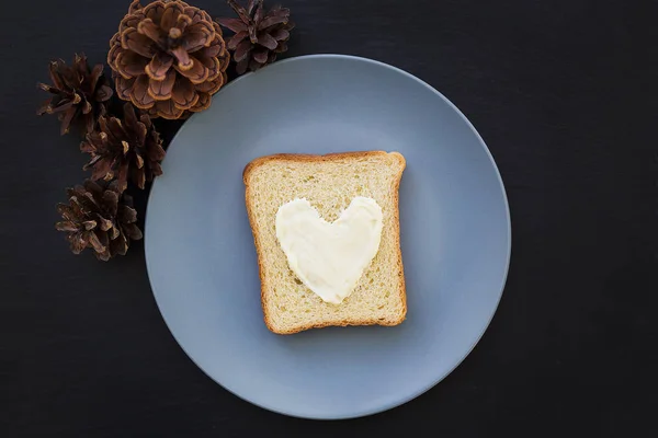 Бутерброд на завтрак в виде сердца с сыром на голубой тарелке и черным фоном с конусами — стоковое фото