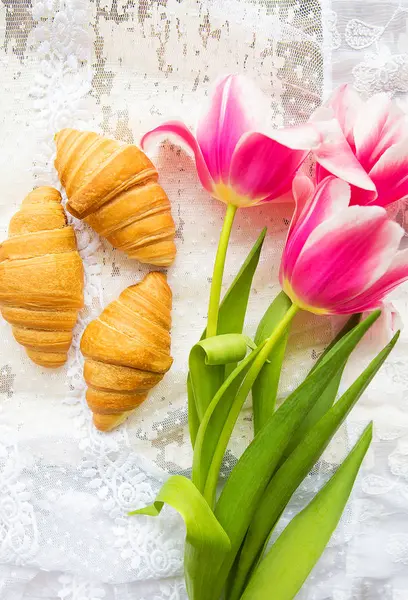 Drei Croissants und leuchtend rosa Tulpen auf Spitzentischdecke — Stockfoto