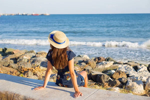 Beautiful girl in a hat sits near the sea on stones — Stock Photo, Image