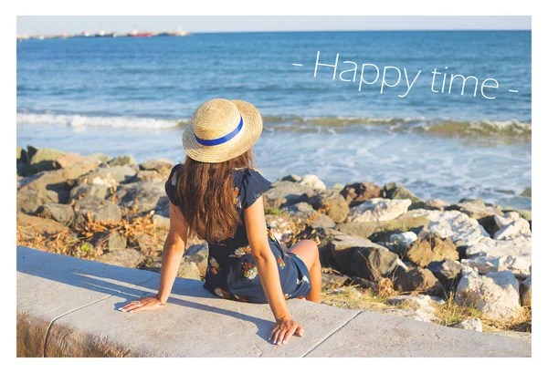 Krásná dívka v klobouku sedí u moře na kamenech, nápis šťastných časů — Stock fotografie