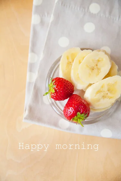 Aardbei en banaan op een glazen plaat op een houten achtergrond, belettering vrolijke ochtend — Stockfoto