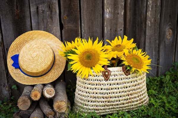 Grumes, chapeau, un bouquet de tournesols dans un sac de paille sont debout près d'une maison en bois . — Photo