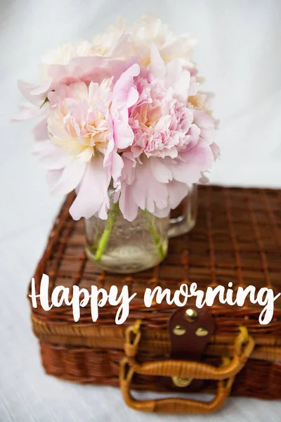 Um buquê muito bonito de peônias rosa em um vaso fica em uma mala de madeira. Inscrição happy morning . — Fotografia de Stock