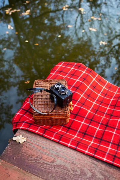 A régi kamera egy bőröndön fekszik egy piros kockás takarón. Fahíd a tó közelében. — Stock Fotó