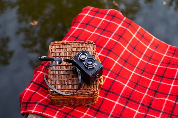 A régi kamera egy bőröndön fekszik egy piros kockás takarón. Fahíd a tó mellett. Közelkép. — Stock Fotó