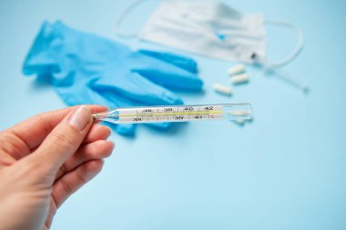 Kızın elinde hapların ve eldivenlerin arka planında termometre var. Tek kullanımlık tıbbi maske ağız ve burnu haplar ve termometreyle mavi arka planda kapatır. Yeni virüs COVID-19.