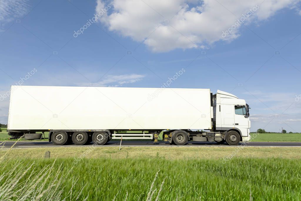 White transport truck driving on the road, green meadow with blue sky