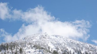 Mavi gökyüzüne karşı karlı dağ zirvelerinde hızlı hareket eden bulutlar.