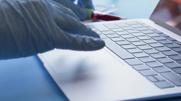 医疗实验室技术员戴着防护乳胶手套 拿着装有血样的试管 用手提电脑输入病人信息 — 图库视频影像