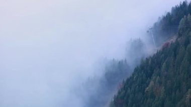 Cable car gondolas moving across the Alps mountain hill from misty cloud to the top. Autumn  misty morning view.  Picturesque traveling, seasonal, cloudy and misty weather condition background scene.