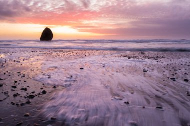 Gold Beach Oregon 'da gün batımı sahnesi. Renkli bulutlar ve dalgalar ıslak kum ve kayalara yansıyan renkler ekliyor.