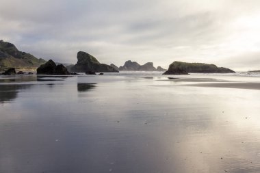 Islak kum Güney Oregon sahilindeki Myers Creek Sahili 'ndeki kayaları ve bulutları yansıtıyor.