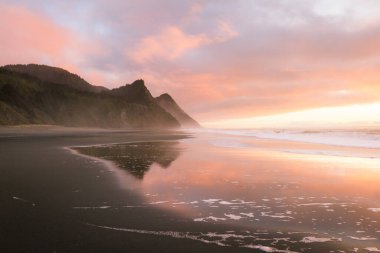 Oregon sahilindeki muhteşem günbatımı sahnesi ıslak kumlara, dalgalara ve Oregon 'un güney sahillerindeki Cape Sebastian tepelerine yansıyan renkli bulutlarla birlikte.