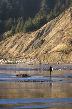 Güneş ışığı arkadaki dağı aydınlatırken kumda sörf tahtası taşıyan bir adam kuma parlak bir yansıma ekliyor.