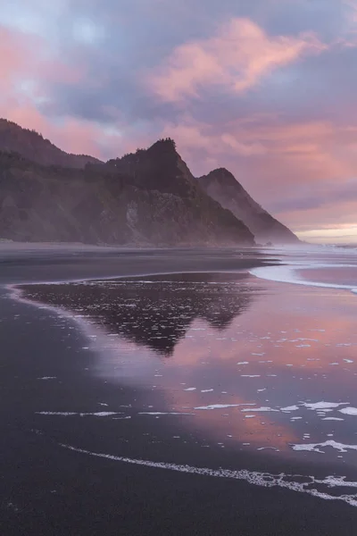 Gorgeous sunset scene in the Oregon coast with colorful clouds reflecting on the wet sand and waves and the hills of Cape Sebastian in the southern Oregon coast