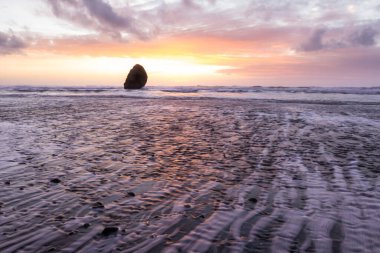 Gold Beach Oregon 'da gün batımı sahnesi. Renkli bulutlar ve dalgalar ıslak kum ve kayalara yansıyan renkler ekliyor.