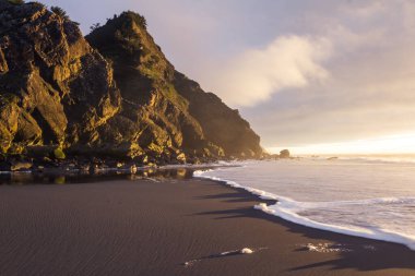 Güneş ufkun altına batarken Güney Oregon sahillerindeki manzaraya parlaklık ve renk katarken, altın ışık Cape Sebastian 'daki kayalara yansıyor.