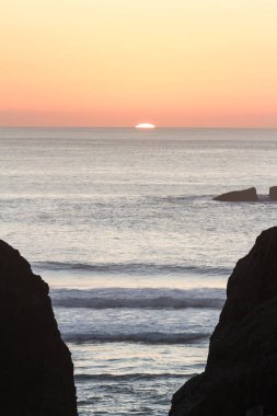 Ufkun ardında parlak turuncu güneş, Bandon Oregon 'daki Face Rock State Park' taki kaya özelliklerini siluete dökerek gökyüzünde ılık tonlar bırakıyor.