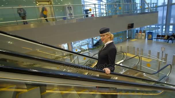 Блондинка-стюардесса поднимается на эскалаторе с чемоданом в аэропорту — стоковое видео