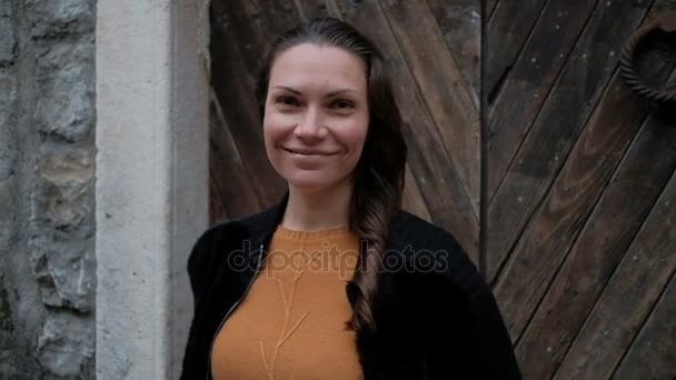 Молодая женщина в оранжевом свитере стоит у деревянной двери и улыбается . — стоковое видео