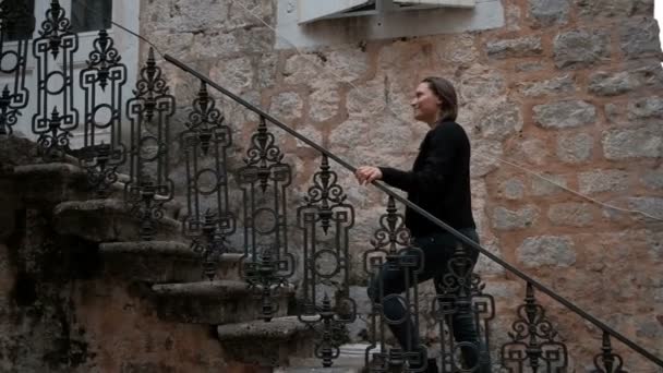 Νεαρή γυναίκα με σκισμένα τζιν επάνω της μια μεταλλική σκάλα από το πέτρινο σπίτι. — Αρχείο Βίντεο