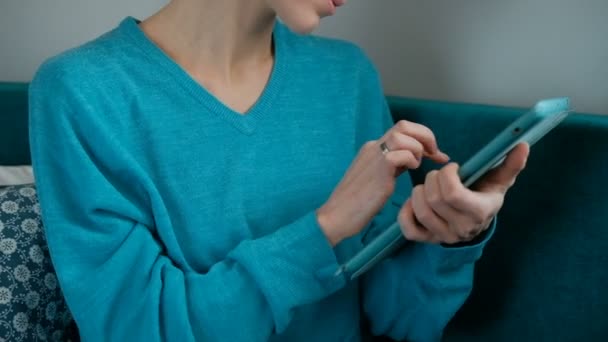 Junge Frau im türkisfarbenen Pullover blättert Tablette auf einer grünen Couch. — Stockvideo