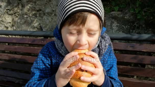 Kleiner Junge genießt es, große Croissants auf Bank im Freien zu essen. — Stockvideo