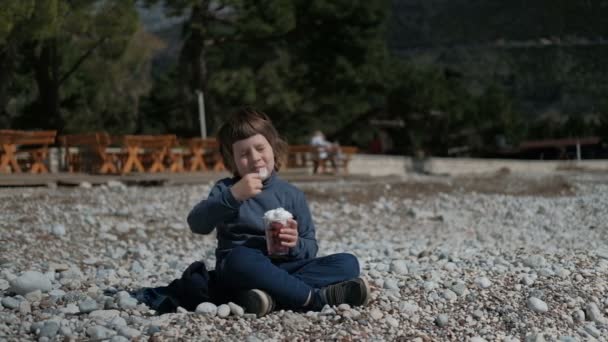 Un niño está sentado en las rocas y come crema con placer. . — Vídeo de stock