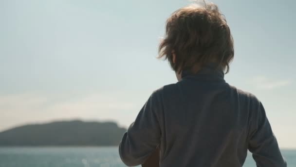 De jongen staat met zijn rug aan de kust en zet op een grote hoed. — Stockvideo