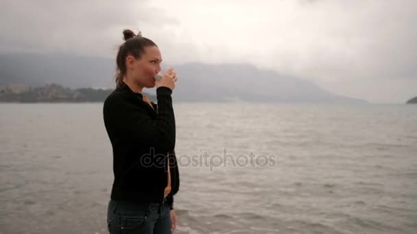 Frau trinkt Kaffee im Stehen am Seeufer, Kamera geht gegen den Uhrzeigersinn. — Stockvideo