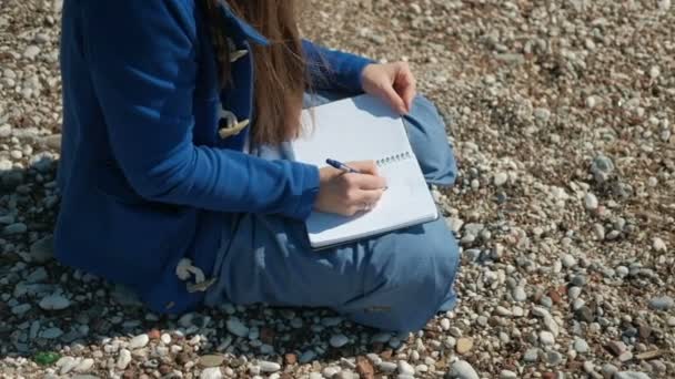 Una mujer está sentada en una playa pedregosa y escribiendo un bolígrafo en el cuaderno . — Vídeo de stock