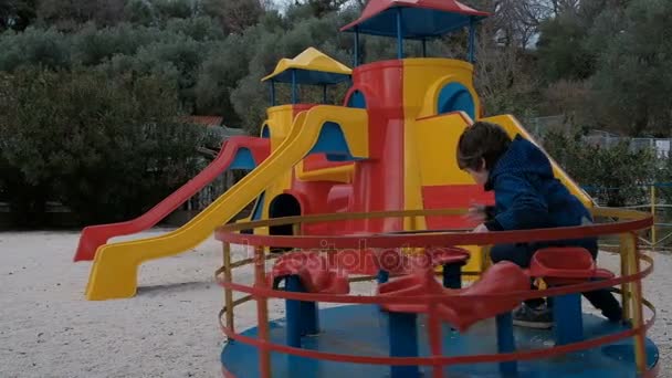 El niño gira rápidamente en el carrusel en el patio de recreo en la primavera — Vídeos de Stock